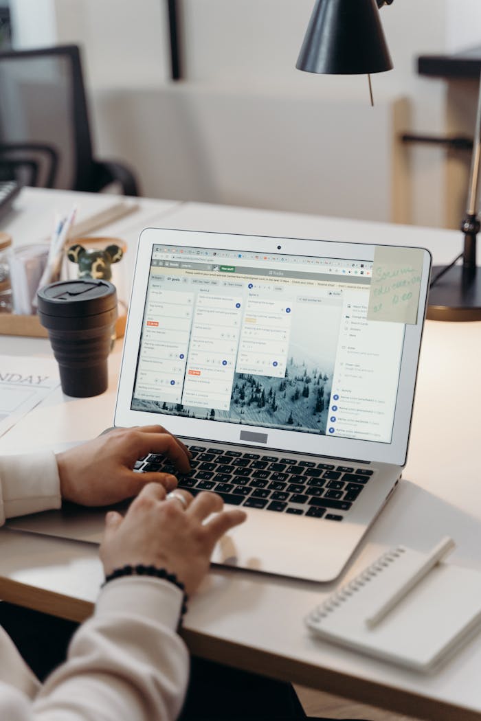 contact-bg Hands typing on a laptop in a modern office setting with notes and task management software visible on the screen.